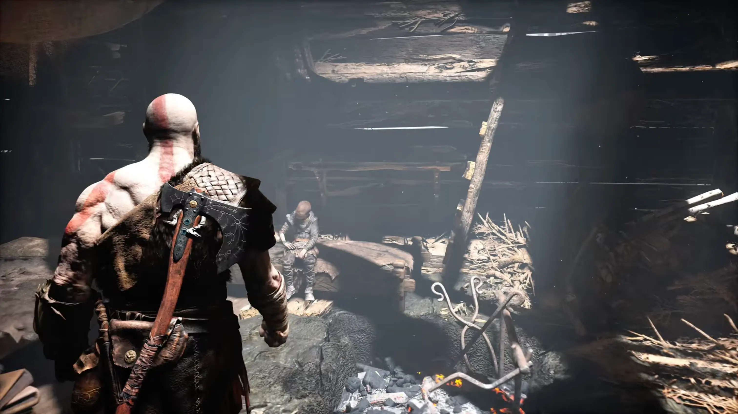 An intimate over-the-shoulder shot of Kratos and Atreus preparing a funeral pyre, showcasing the game's brilliant, unbroken cinematic camera angle.
